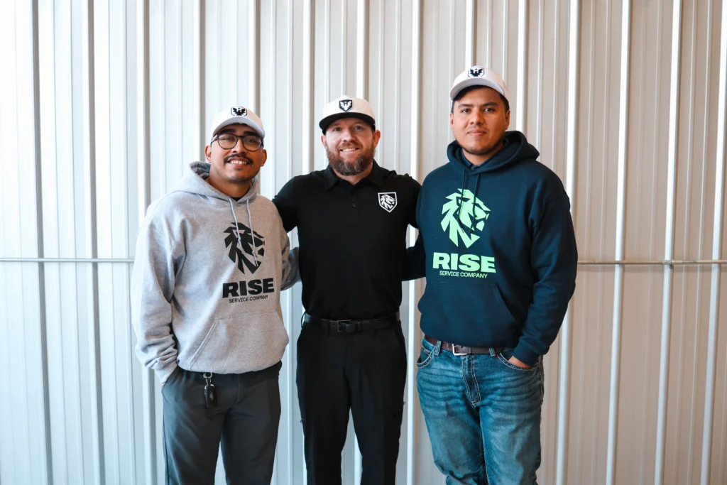 Three male employees of RISE Service Company wearing branded hoodies and hats, smiling in front of a neutral industrial wall.